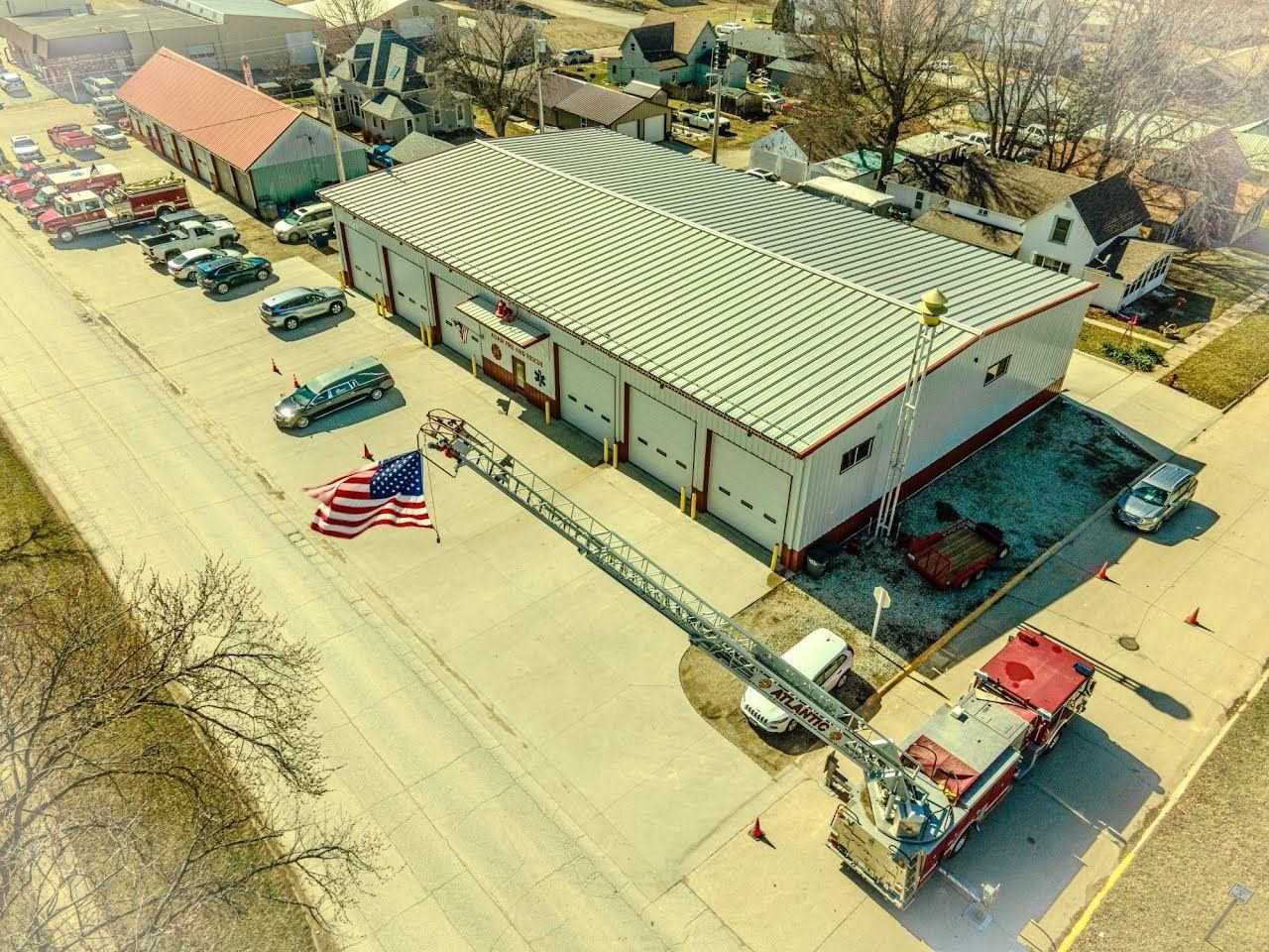 Aerial drone photo of a fire station, ladder truck, American flag, rooftops, and surrounding town streets